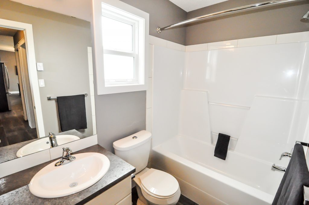 Modern bathroom with a white bathtub, gray walls, and dark towel on a rail. Includes a sink, mirror, window, and dark countertop. Clean and minimalist.