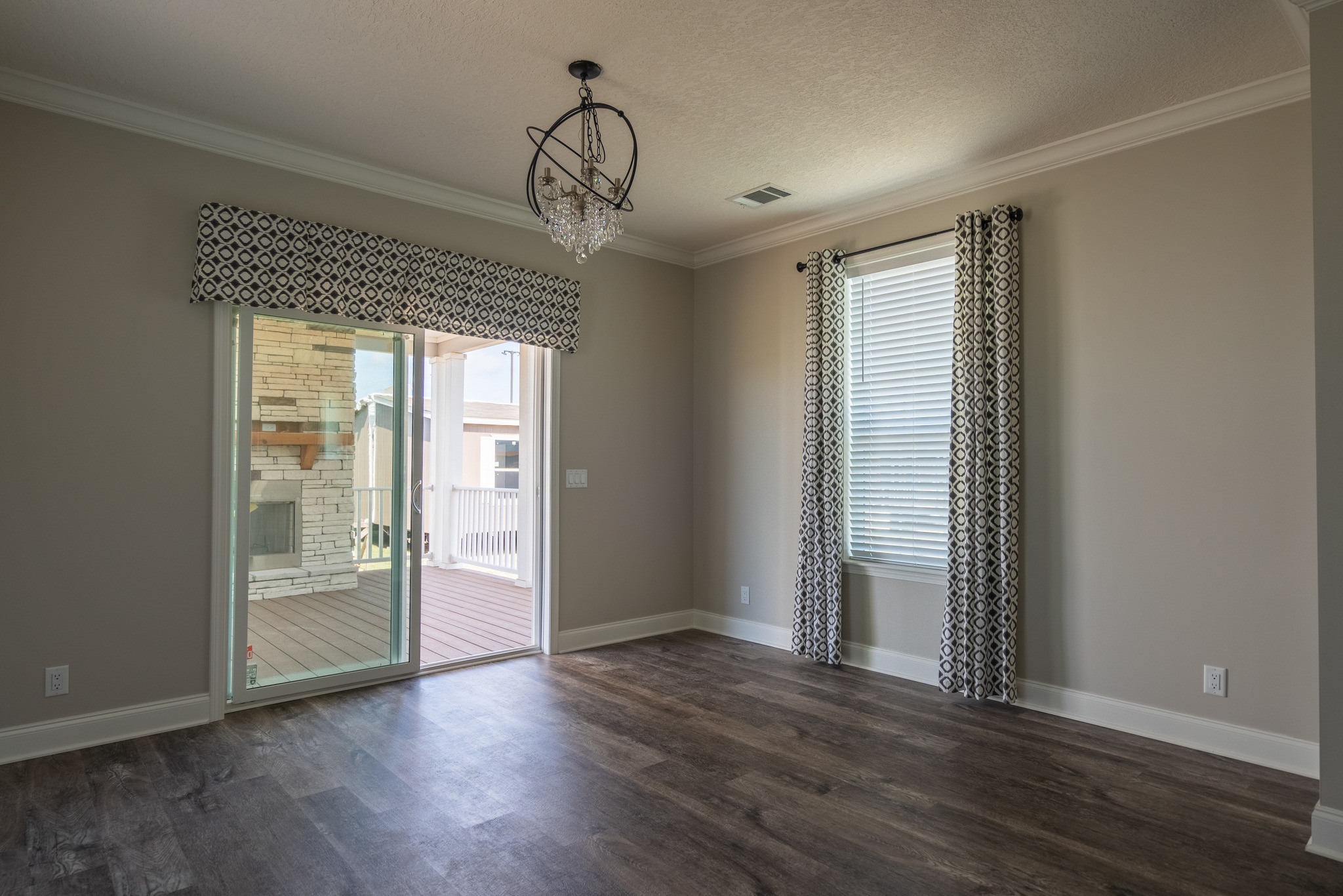 Empty room with wood flooring, beige walls, and a modern chandelier. Large sliding glass door on the left and a window with patterned curtains on the right.