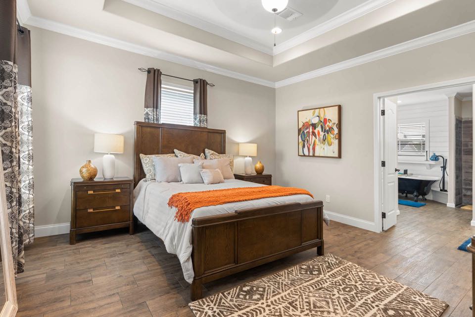 Spacious bedroom with a wooden bed, orange blanket, and cushioned headboard. Warm lighting from bedside lamps, abstract art on the wall, open door to bathroom.
