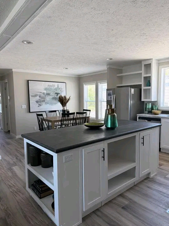 Modern kitchen and dining area with a sleek gray island, green decor accents, abstract wall art, and a wooden dining table. Bright, airy atmosphere.