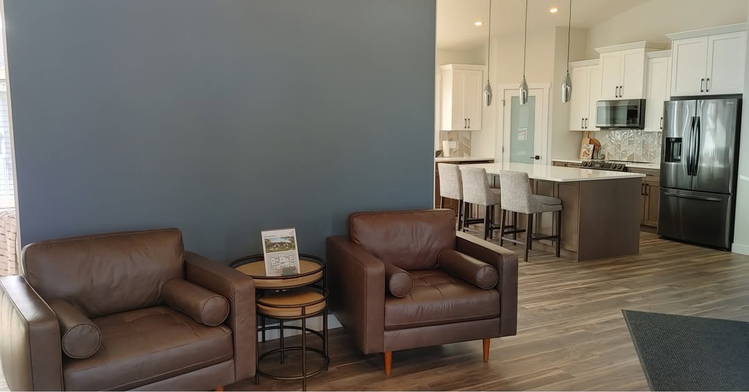 Two brown leather armchairs sit by a dark blue wall with a small round table in between. The modern kitchen features white cabinets and stainless appliances, creating a cozy, inviting atmosphere.