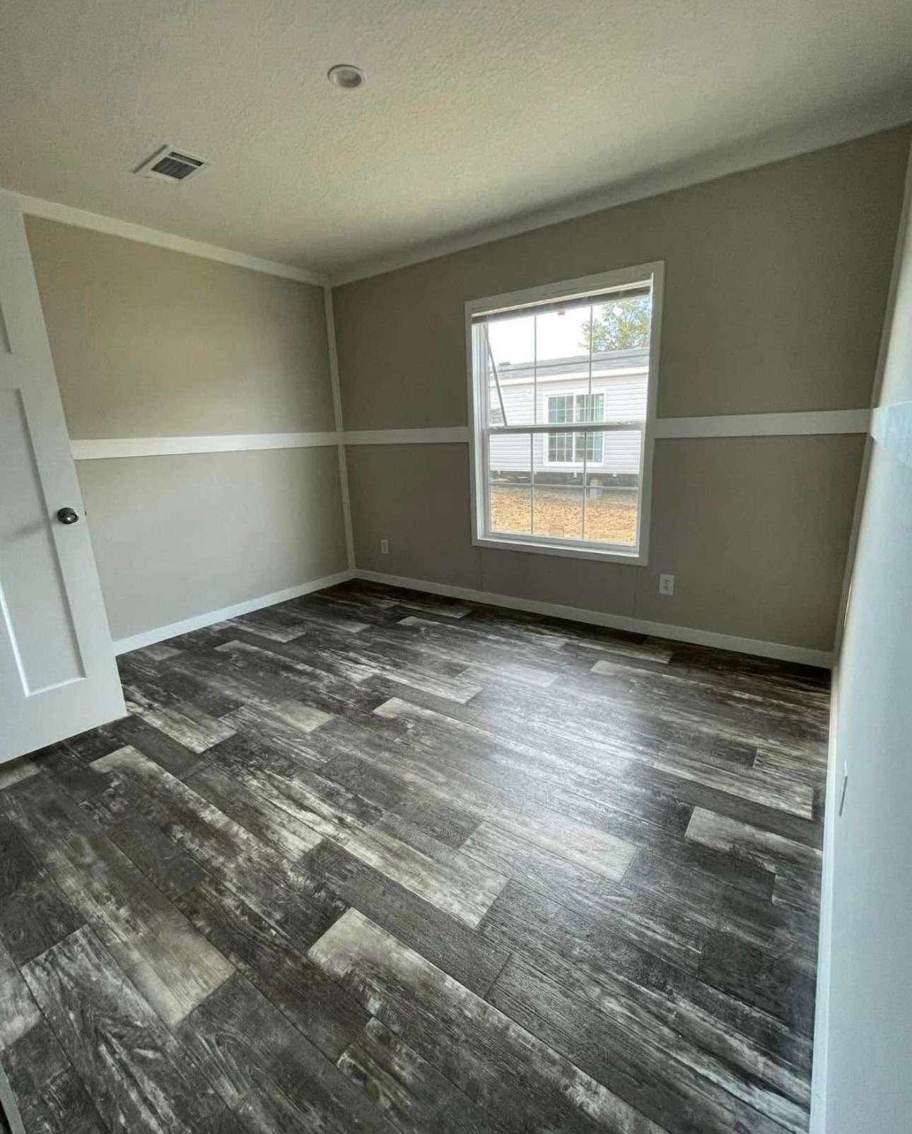 Empty room with gray wood-patterned flooring and beige walls, featuring a large window with a view of another building, creating a calm and spacious atmosphere.
