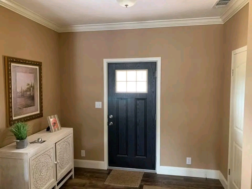 A cozy entryway with warm beige walls and a dark wood door with a window panel. A light wood cabinet holds a plant and picture frames, beside a serene wall painting.