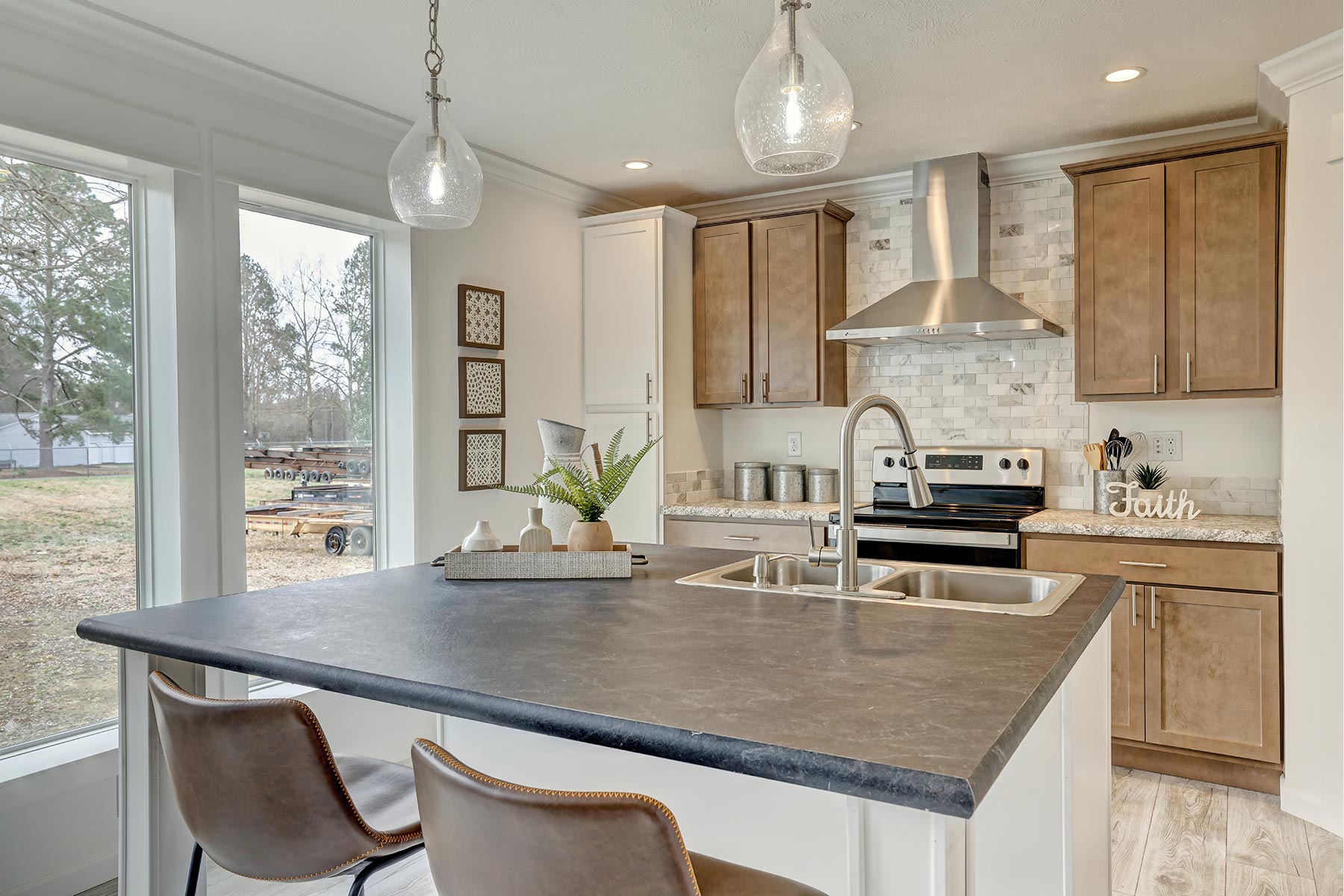 Modern kitchen with a gray countertop island, stainless steel appliances, and light wood cabinetry. Large windows offer a view of a green yard.