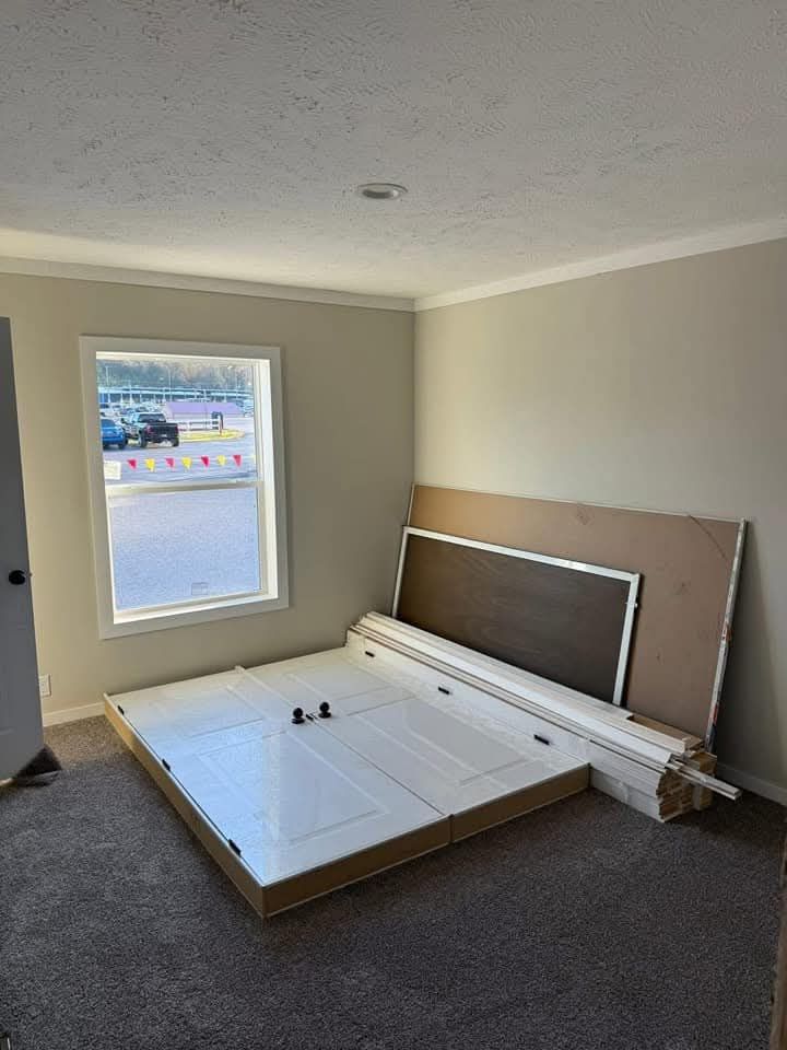 An empty room with beige walls and carpet. A window reveals parked cars outside. Pieces of a disassembled white door and frame are stacked in the corner.