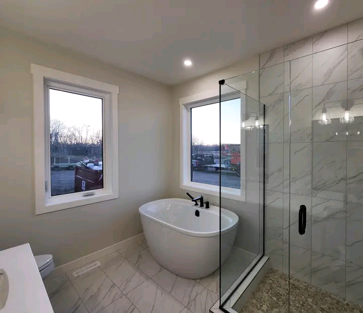 Modern bathroom with a freestanding white tub, black fixtures, large windows, and a glass-enclosed shower. The neutral tones create a serene atmosphere.