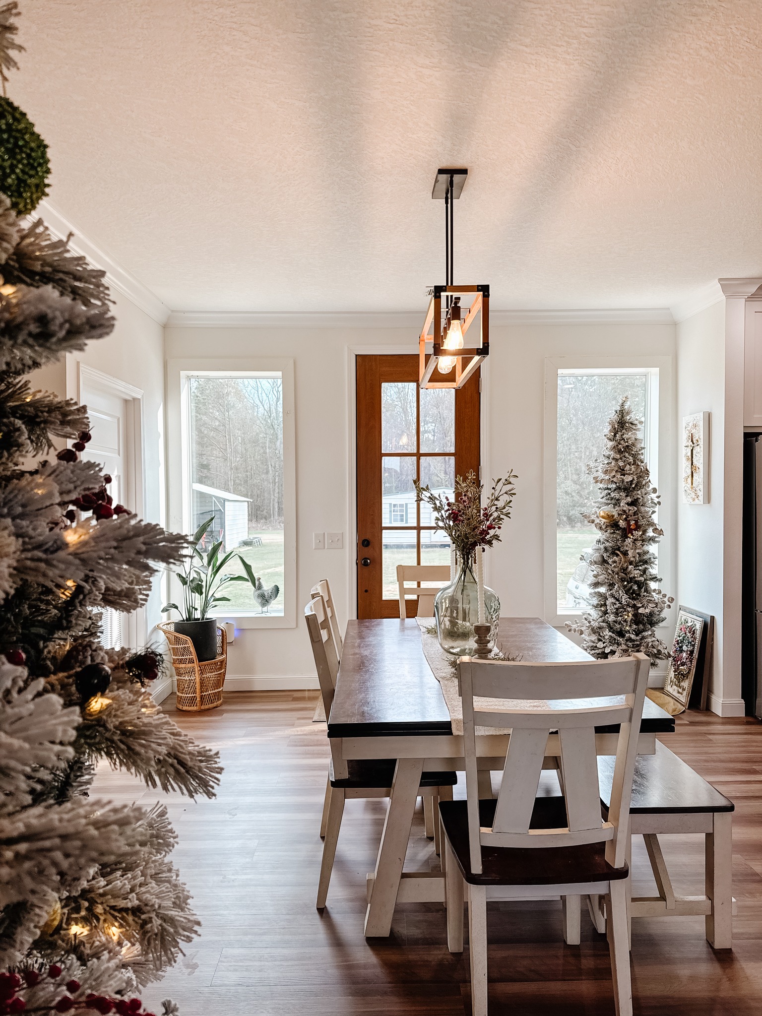 Bright dining area with a rustic wooden table, white chairs, and festive decor. Snow-covered trees and plants frame the sunlit window view. Cozy ambiance.