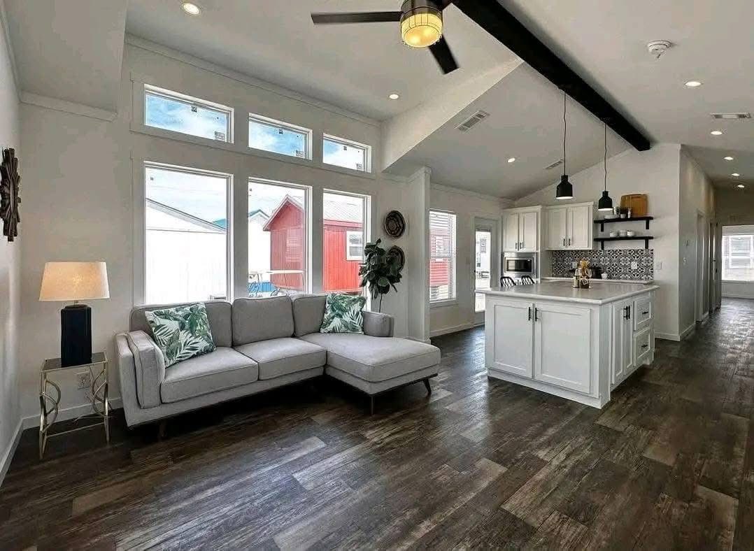 Modern living room with wood floors, gray L-shaped sofa, and large windows. Bright kitchen has white cabinets and island, pendant lights, and a ceiling fan. Cozy and airy feel.