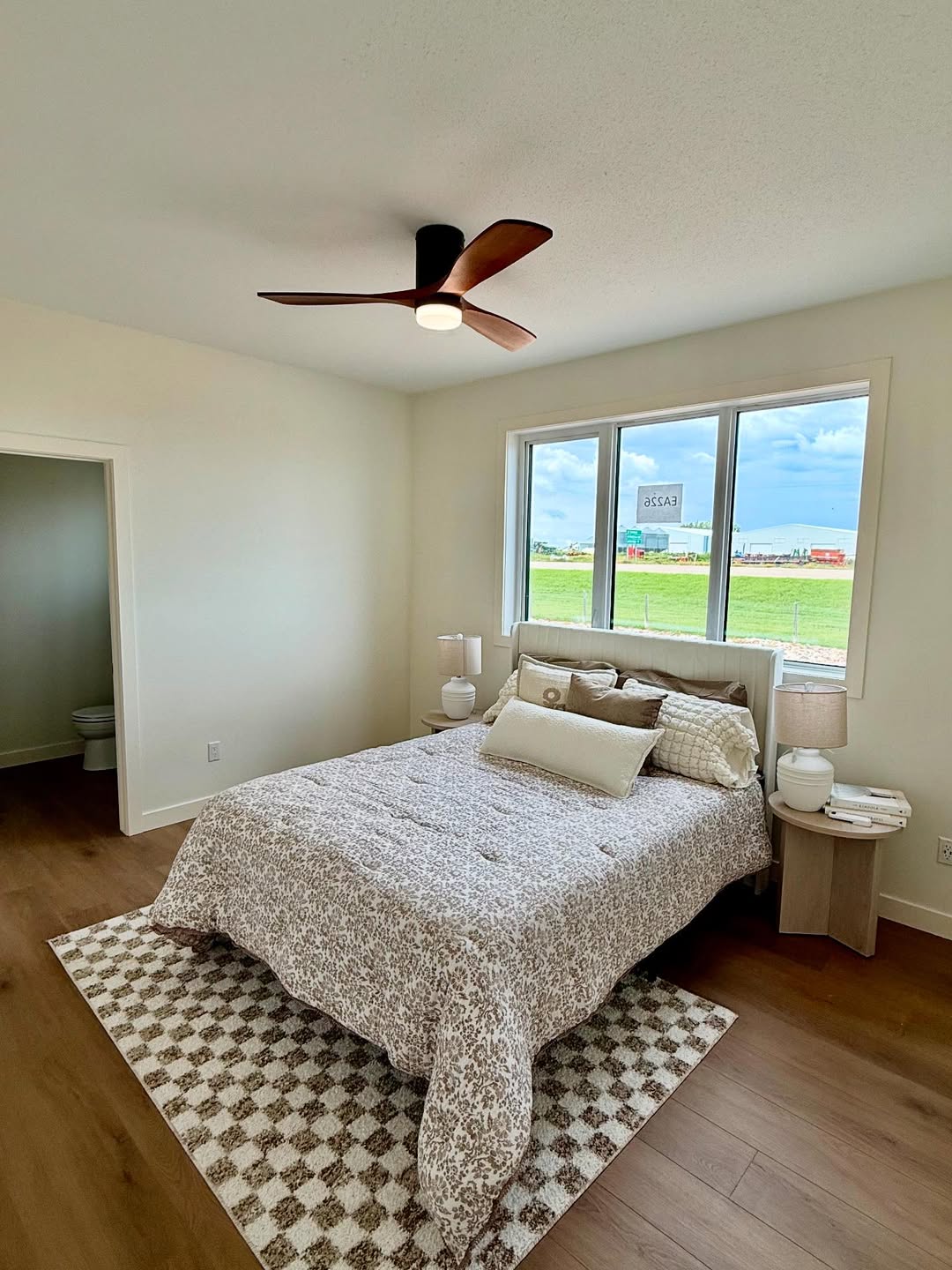 Bright bedroom with a neatly made bed covered in a patterned comforter, flanked by wooden nightstands with lamps. Large window reveals a green field.