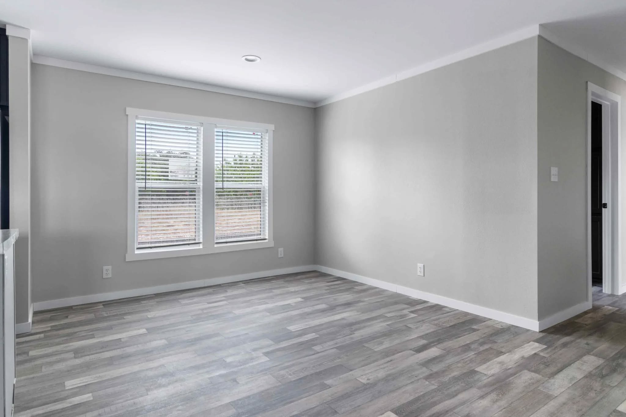A minimalist, empty room with pale gray walls and wood-patterned flooring. Sunlight filters through two windows with white blinds, creating a serene atmosphere.