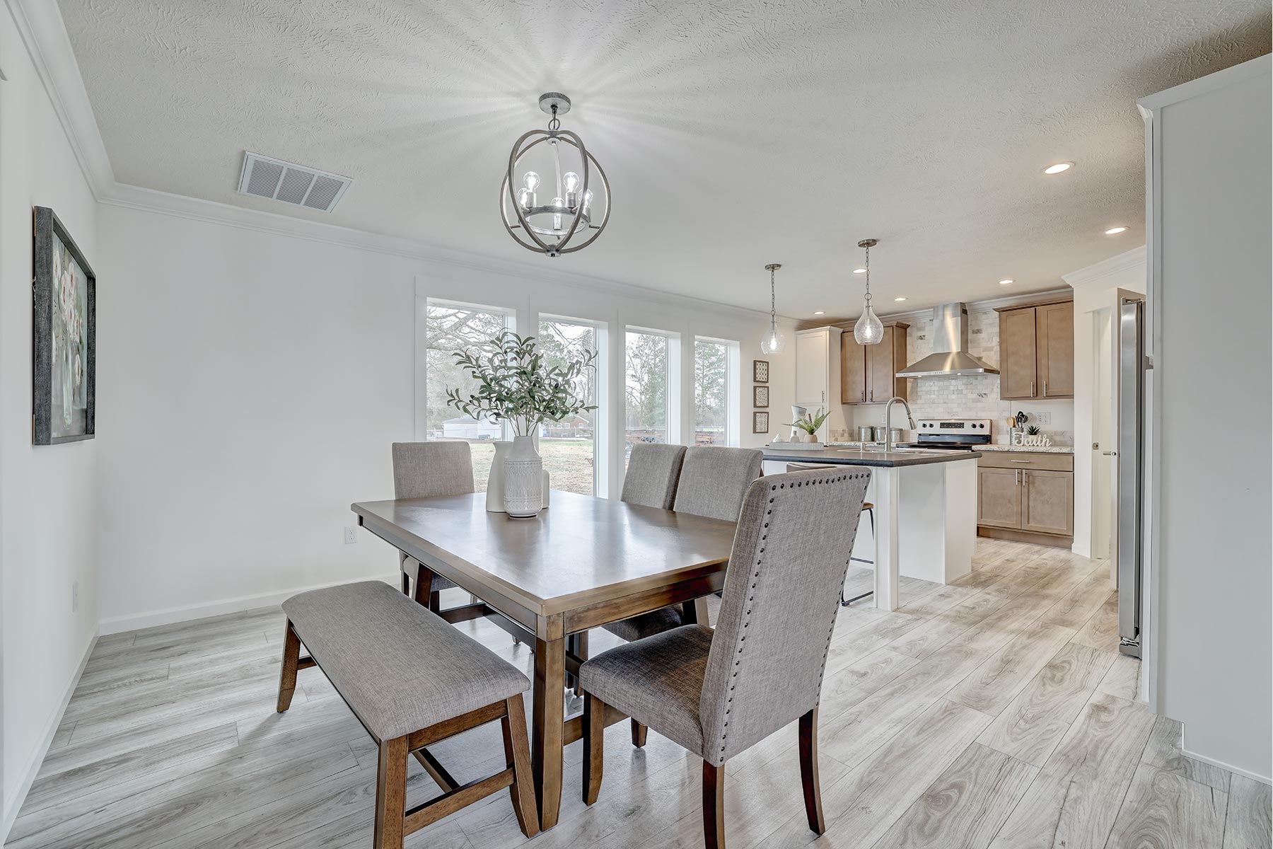 Modern dining area with a wooden table, gray upholstered chairs, and a bench. Pendant lighting, light wood flooring, and open kitchen in the background convey a cozy, welcoming atmosphere.