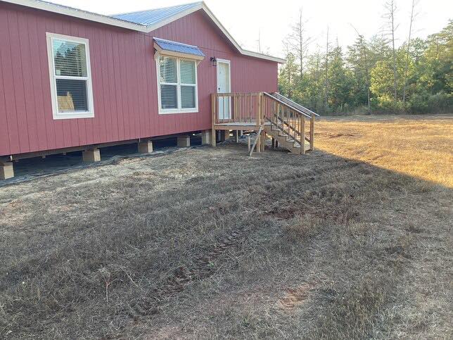 A small red house with white trim sits on a dry grassy plot. Wooden steps lead to the door. Sparse trees line the background under a clear sky.