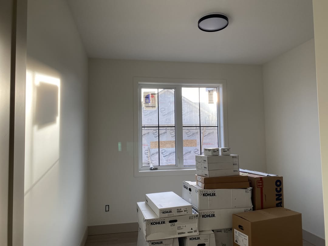 A small, empty room with light-colored walls. Sunlight filters through a window, casting a glow. Boxes labeled "Kohler" are stacked in the corner, suggesting renovation.