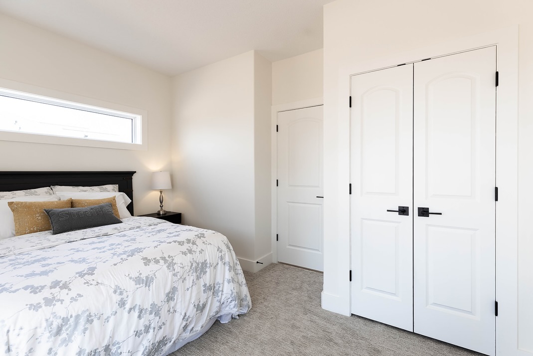 Bright bedroom with white walls and cozy carpet features a double bed with floral bedding, bedside lamp, and large white closet doors.