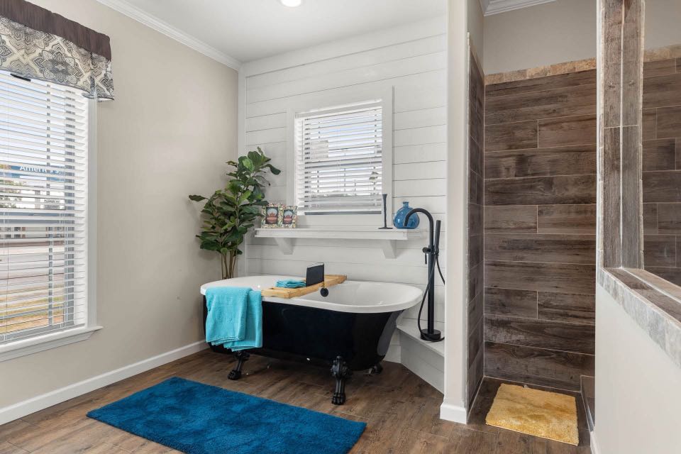 A cozy bathroom with a freestanding black tub, blue towel, and wooden floor. Nearby, a plant and window enhance brightness. Rustic wood accents add warmth.