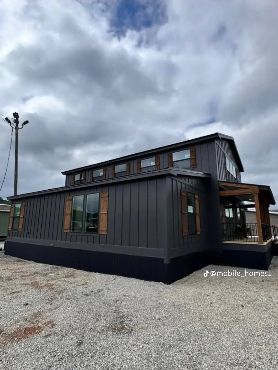 Modern mobile home with dark siding and wooden shutters under a cloudy sky. The structure is angular and sits on a gravel surface, giving a contemporary feel.