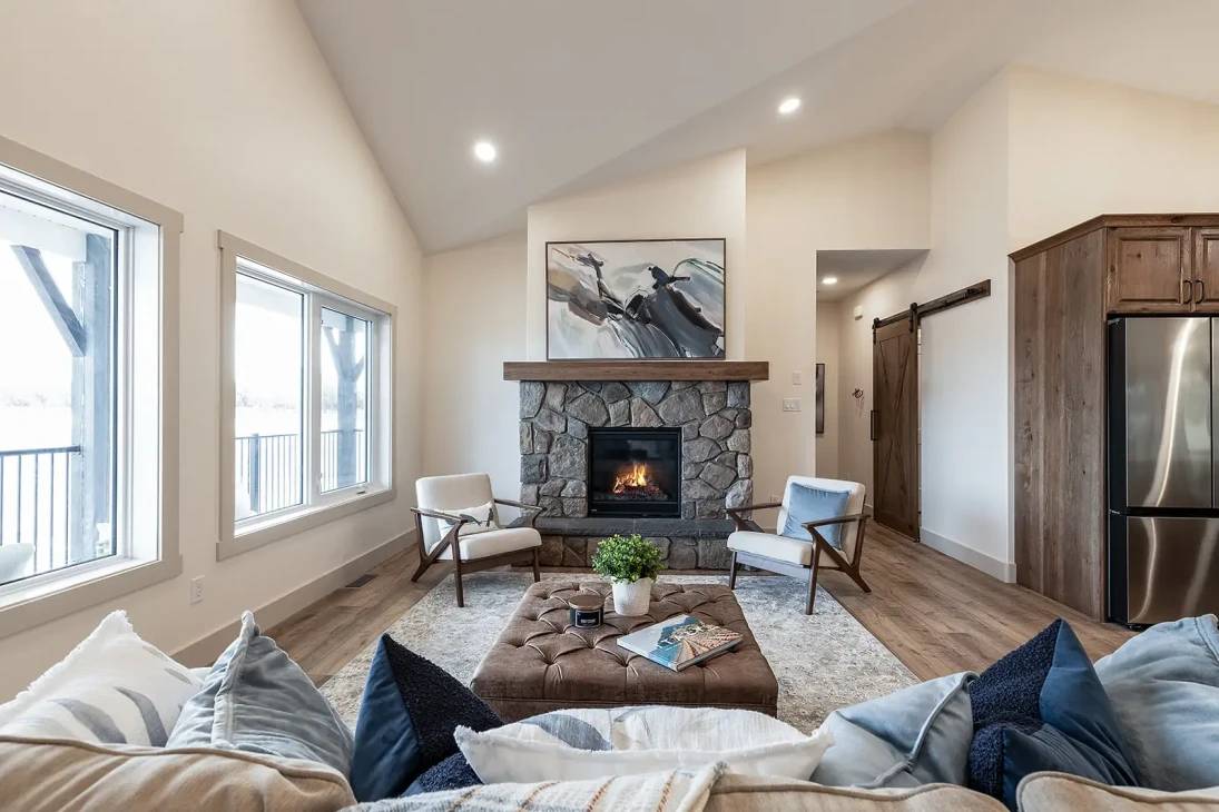 Cozy living room with stone fireplace, abstract art, and comfy chairs. Soft lighting, wooden floors, and a large window create a warm, inviting atmosphere.