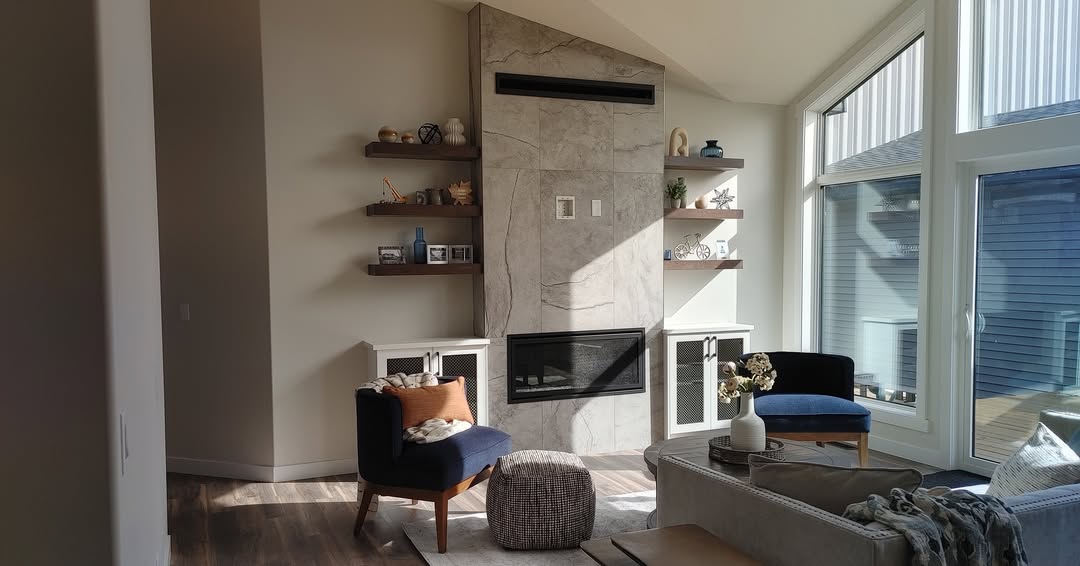 Bright living room with large windows, a modern stone fireplace, and wooden shelves. Cozy chairs, a couch, and decor create a warm, inviting atmosphere.