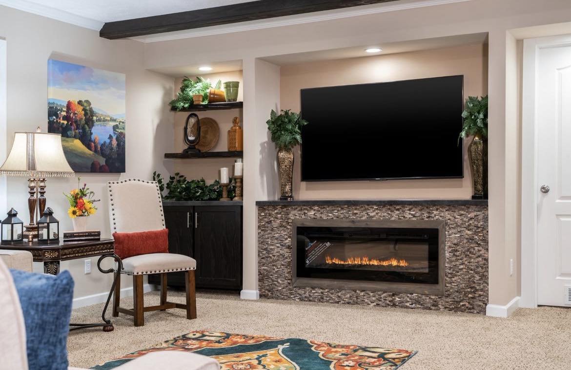A cozy living room with beige walls, a large flat-screen TV, and a modern electric fireplace. Shelves hold vases and decor. A colorful painting and plush seating add warmth.
