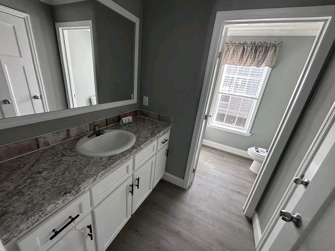 Modern bathroom with gray walls, a marble countertop, and white cabinets under a rectangular mirror. An open door reveals a bright toilet area with a window.