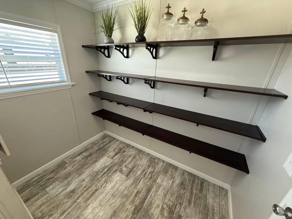 A small, bright pantry with several dark wooden shelves on white walls. The top shelf holds decorative jars and green plants, creating a minimalist, organized feel.