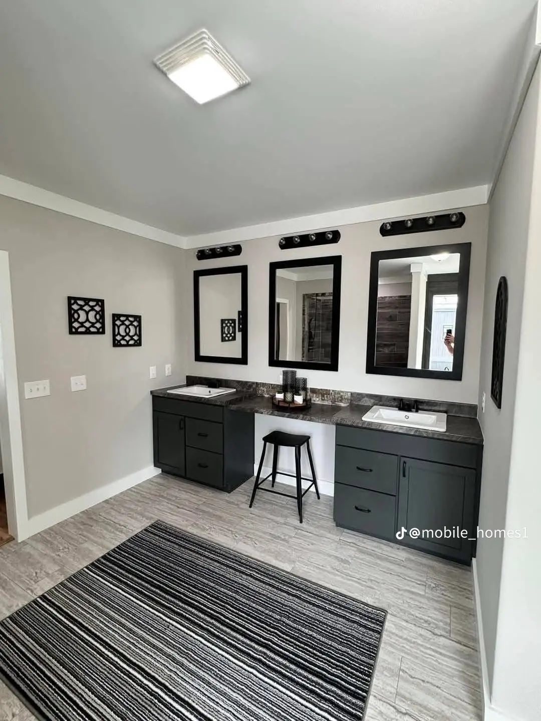Spacious modern bathroom with dual sinks and dark cabinets. Three black-framed mirrors above, a black stool centered, and a striped rug on the tiled floor.