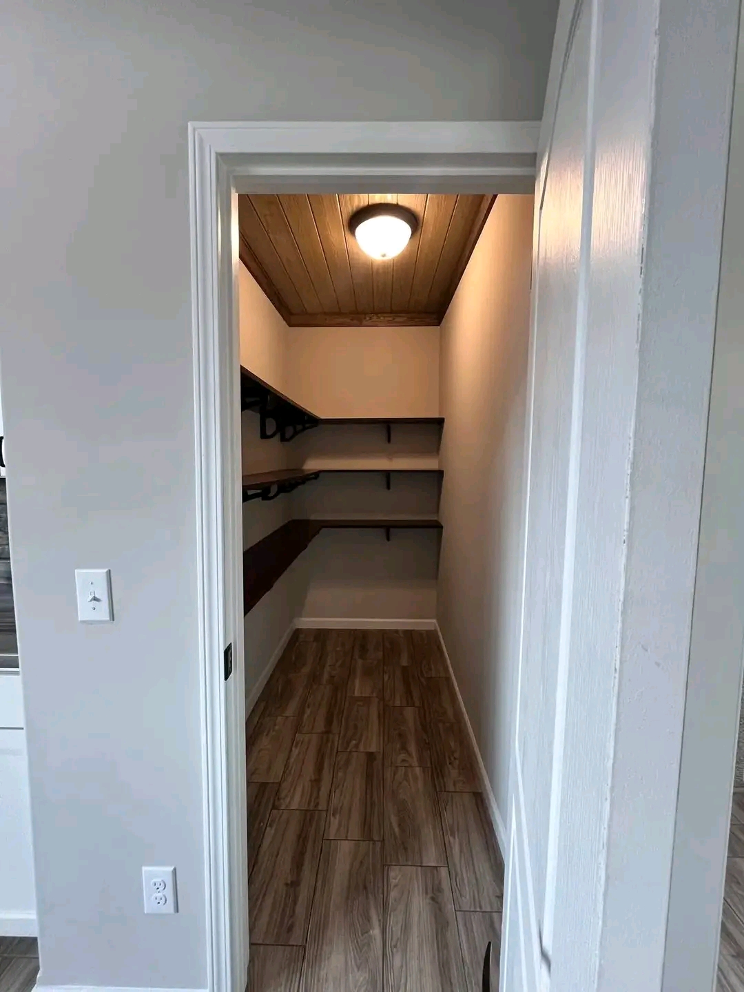 A narrow pantry with wooden shelves on both sides is lit by a ceiling light. The walls are light-colored, and the floor features wood-like tiles.