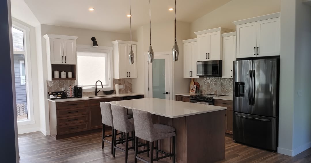 Modern kitchen with white cabinets, marble countertops, and stainless steel appliances. An island with chairs is in the center, under pendant lights.
