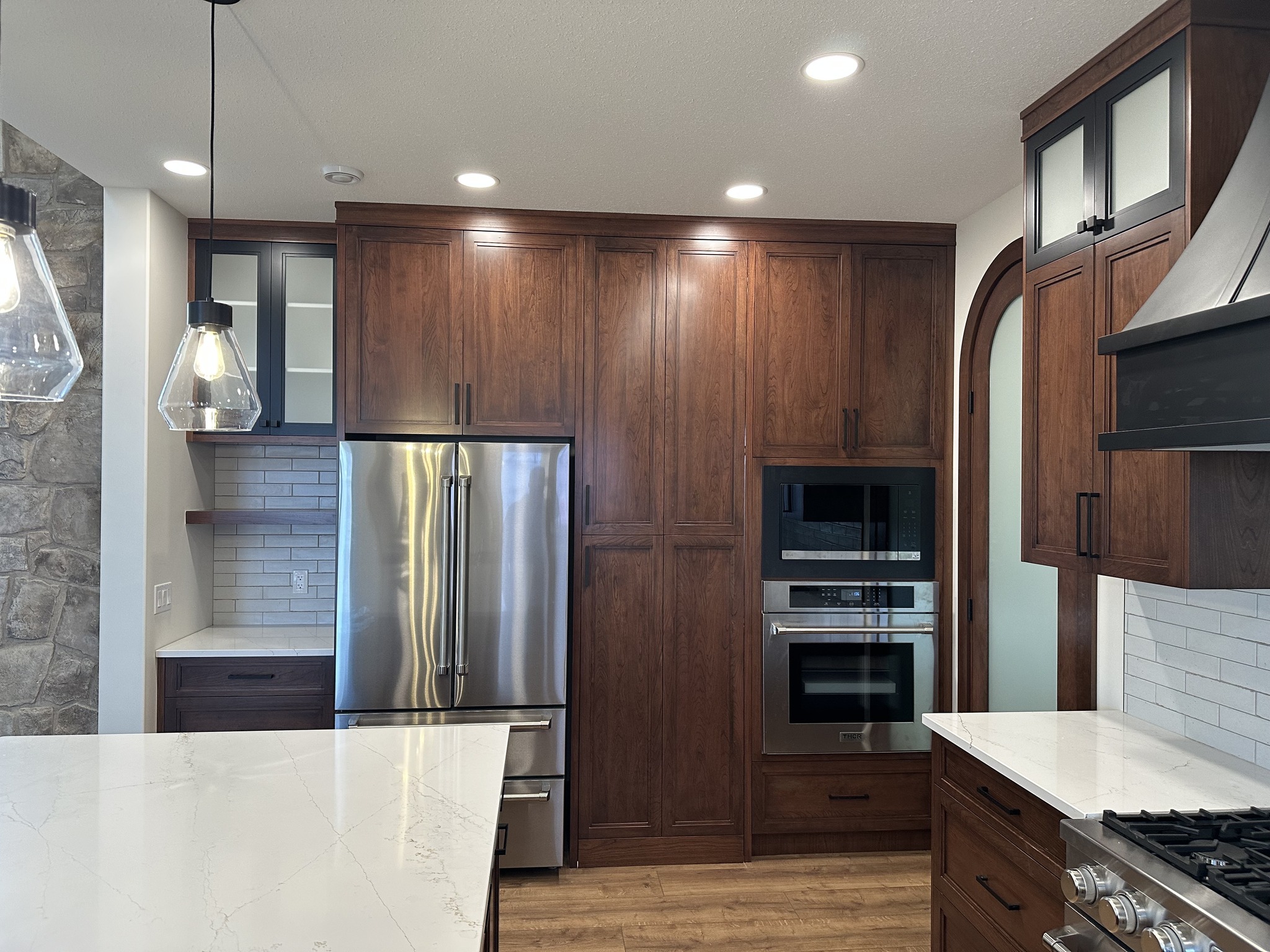 Modern kitchen with wooden cabinets, stainless steel fridge, double oven, and marble countertops. Pendant lights and stone accent wall add elegance.