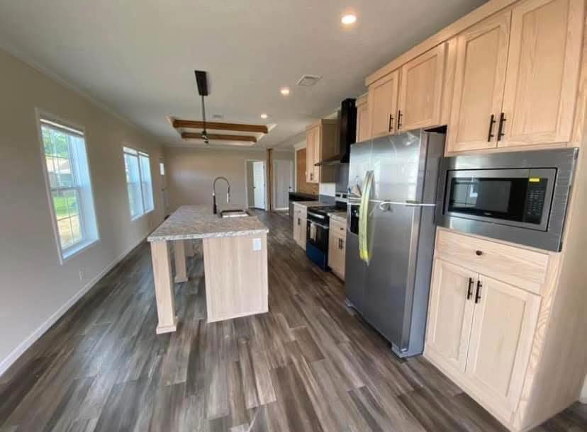 Modern kitchen with light wood cabinets, stainless steel appliances, and a marble island. Dark wood floor adds warmth, large windows offer natural light.