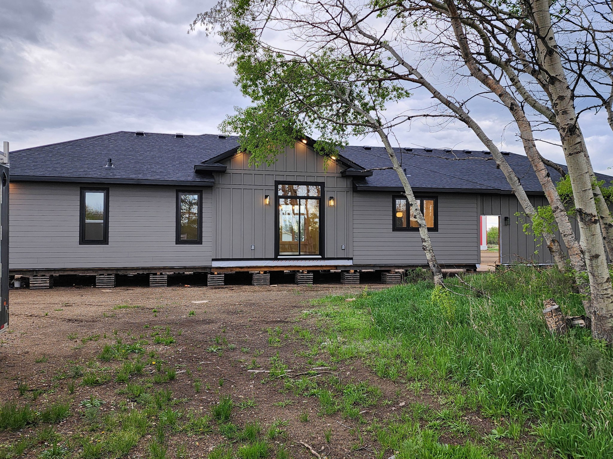 A modern gray house on raised blocks, framed by sparse trees and green grass. Overcast sky sets a calm, neutral tone. Construction site feel.