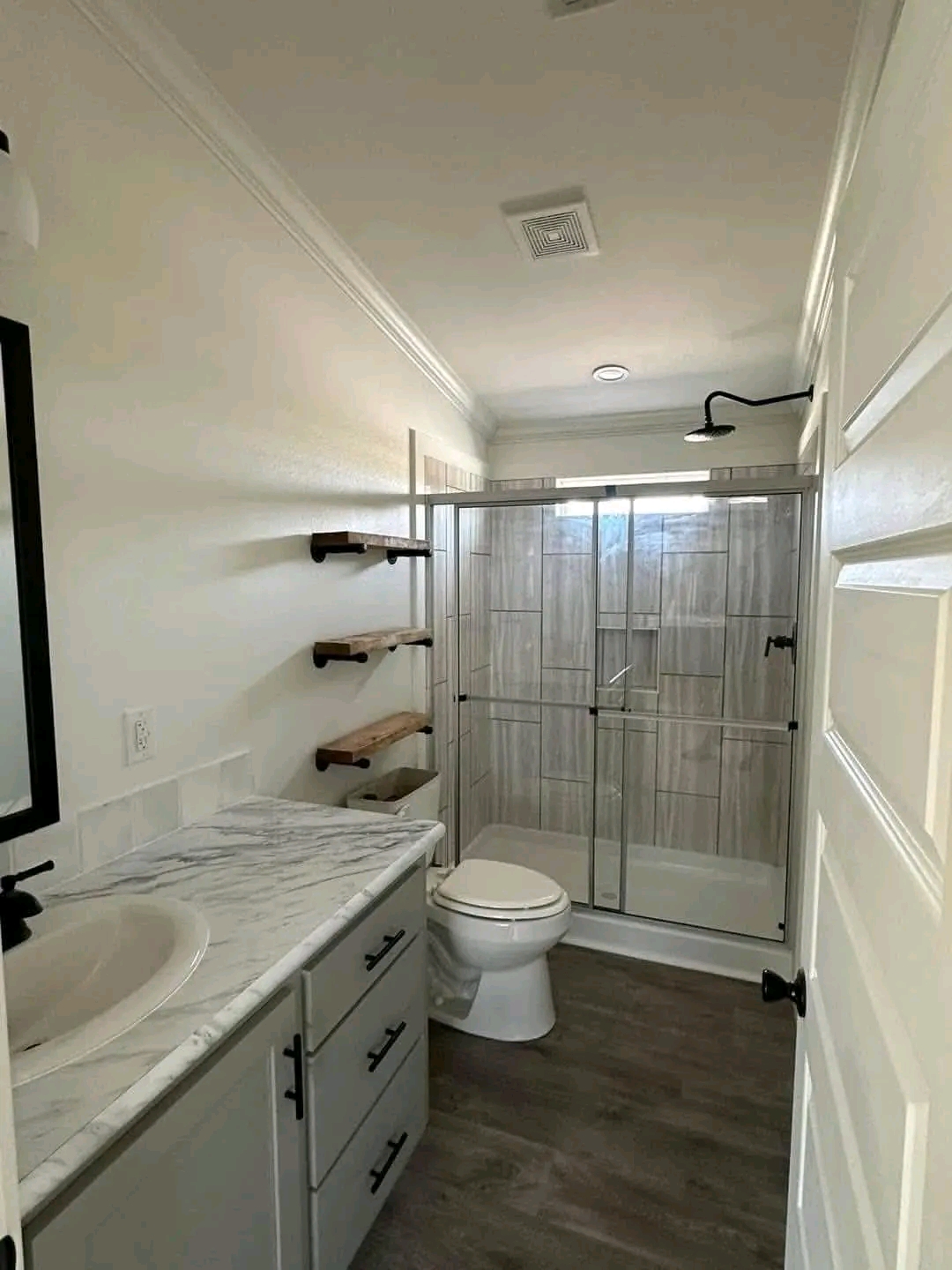 Modern bathroom with a marble countertop, white sink, and storage drawers. Wooden shelves on the wall, toilet, and glass-enclosed shower. Bright, clean, and minimalistic.