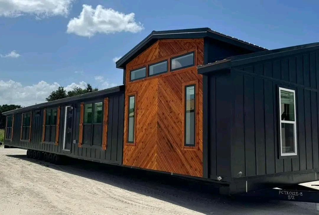 A modern, mobile home with a sleek dark exterior and wooden accents is parked on a dirt road. Large windows allow natural light, creating a welcoming feel.