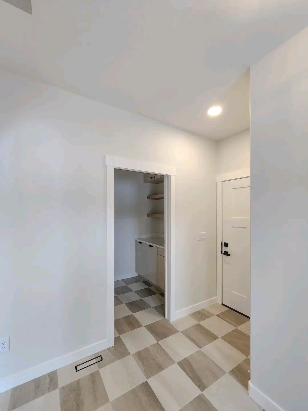 Bright entryway with white walls and checkered beige flooring. A white door with black handle is on the right, and an open doorway leads to a small room with shelves.