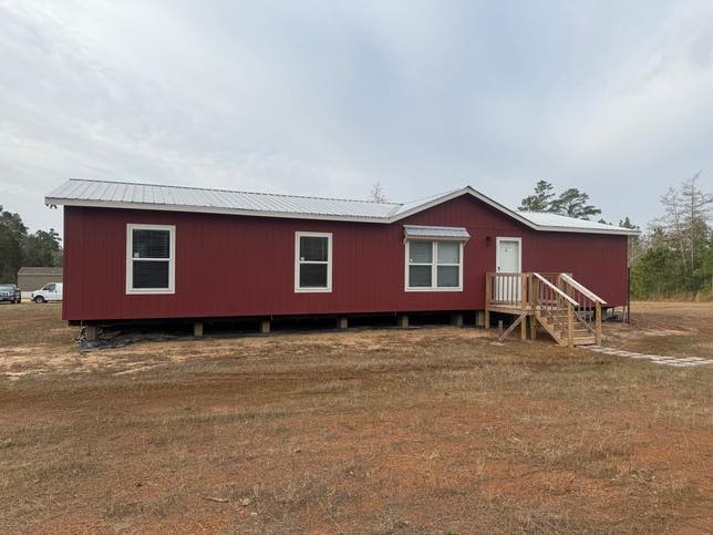 A red, single-story modular home with a white door and windows, set on a grassy field under an overcast sky. Wooden steps lead up to the entrance.