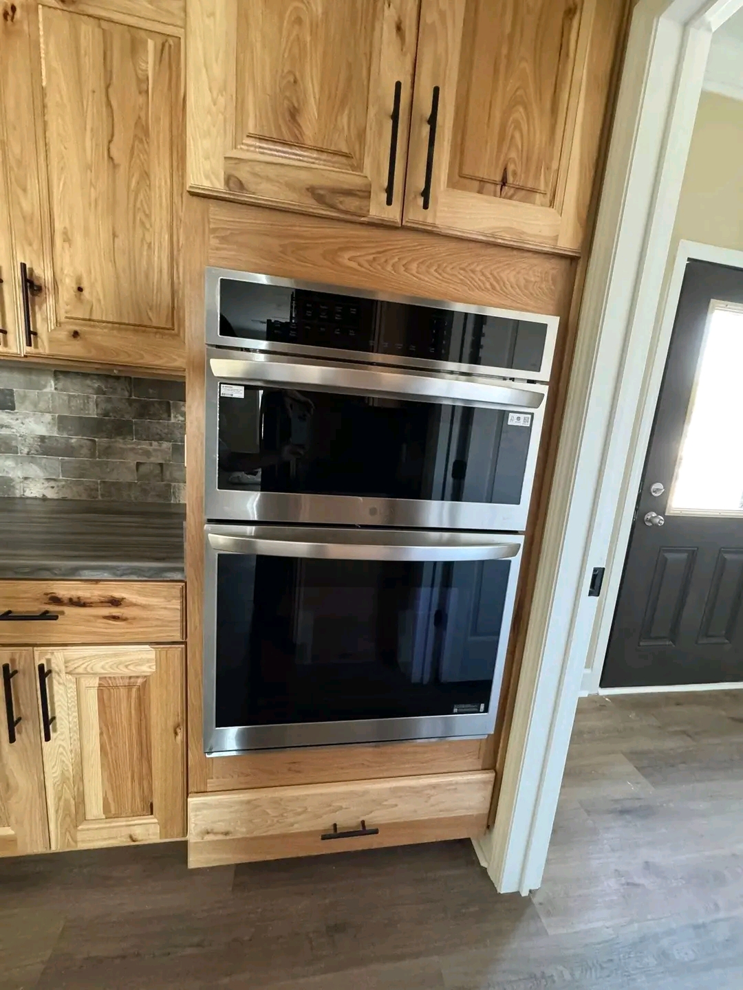Built-in stainless steel double oven framed by light wood cabinets and dark handles. Adjacent is a gray stone backsplash and wooden countertop. Cozy kitchen ambiance.