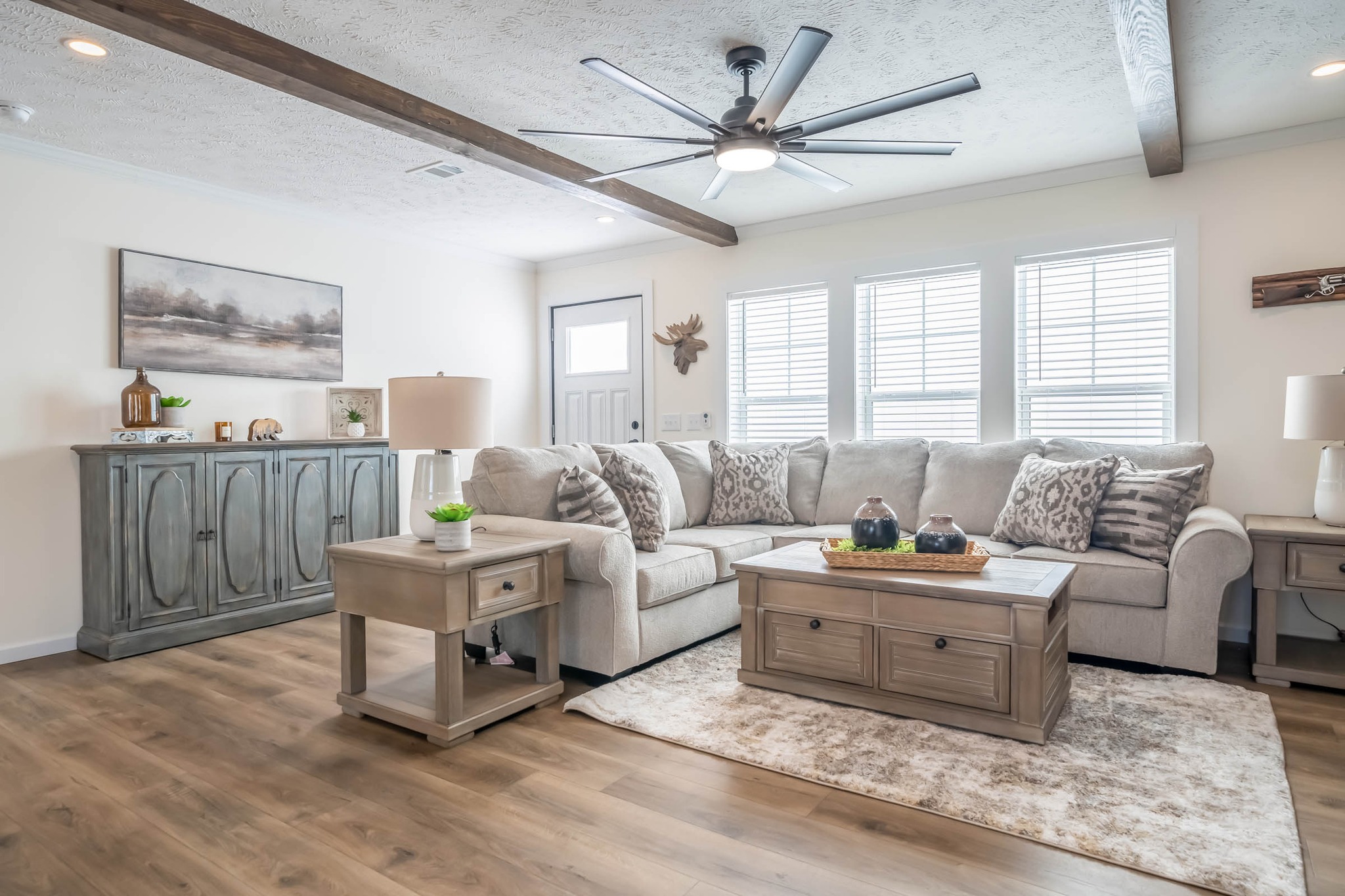 Contemporary living room with a beige sectional sofa, patterned pillows, wooden coffee table, side tables, large ceiling fan, and natural light from windows.