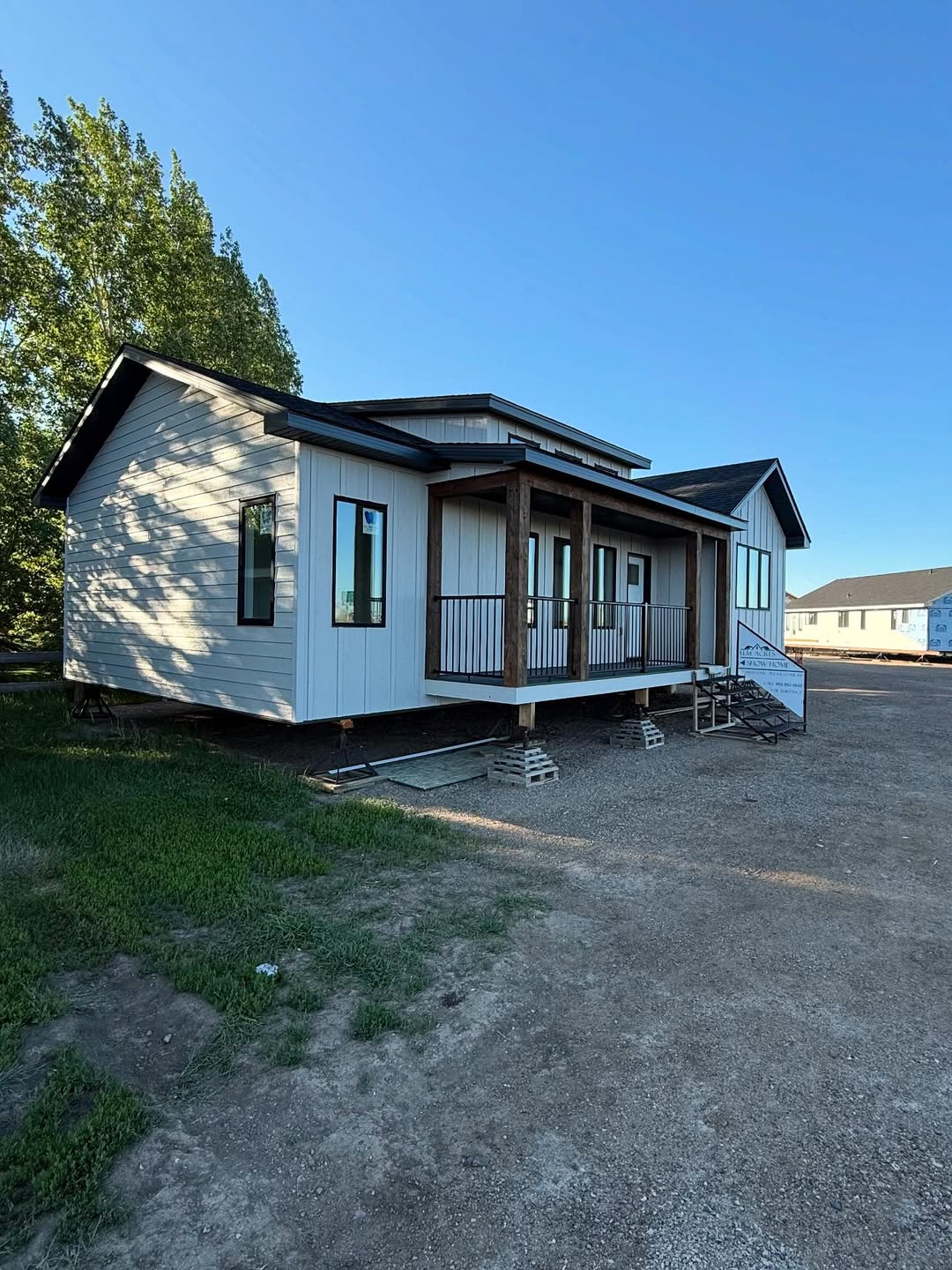 Single-story modular home with a white exterior, dark trim, and a small porch sits on an empty lot with clear blue skies and trees in the background.