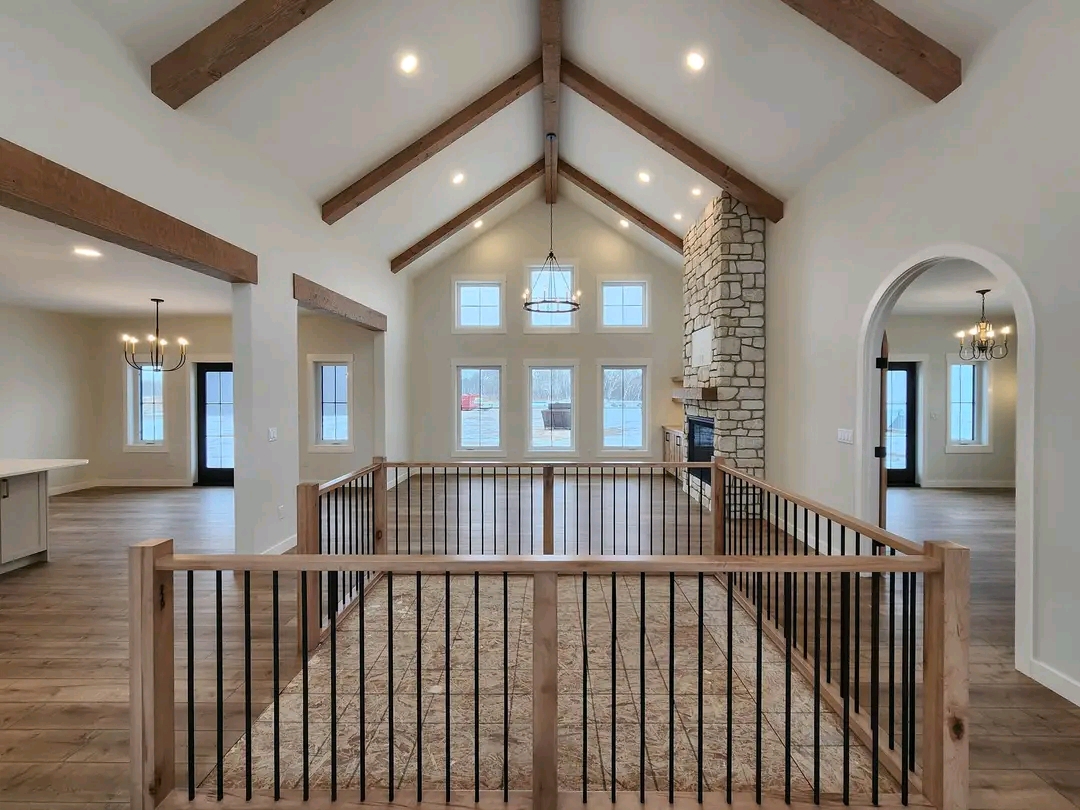 Spacious living room with high vaulted ceiling and wooden beams. Stone fireplace, large windows, and modern chandelier create a warm, inviting atmosphere.