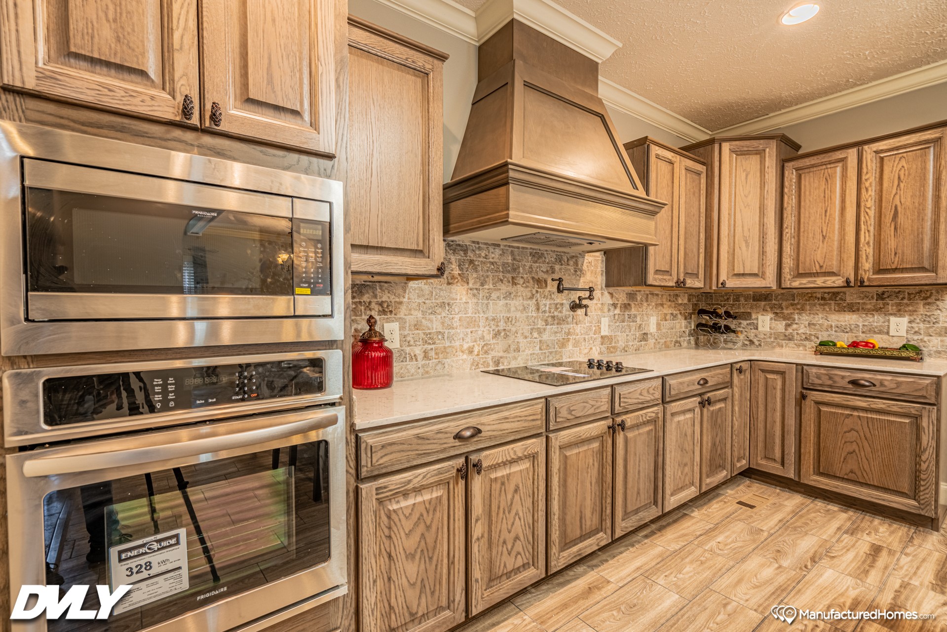 Warm kitchen interior with oak cabinets, stainless steel oven, microwave, and range hood. Brick backsplash and wood floors create a cozy feel.