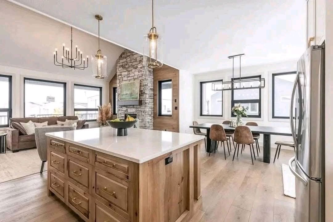 Spacious modern kitchen and living area features a large wooden island with a white countertop. Elegant chandeliers and natural light add warmth.
