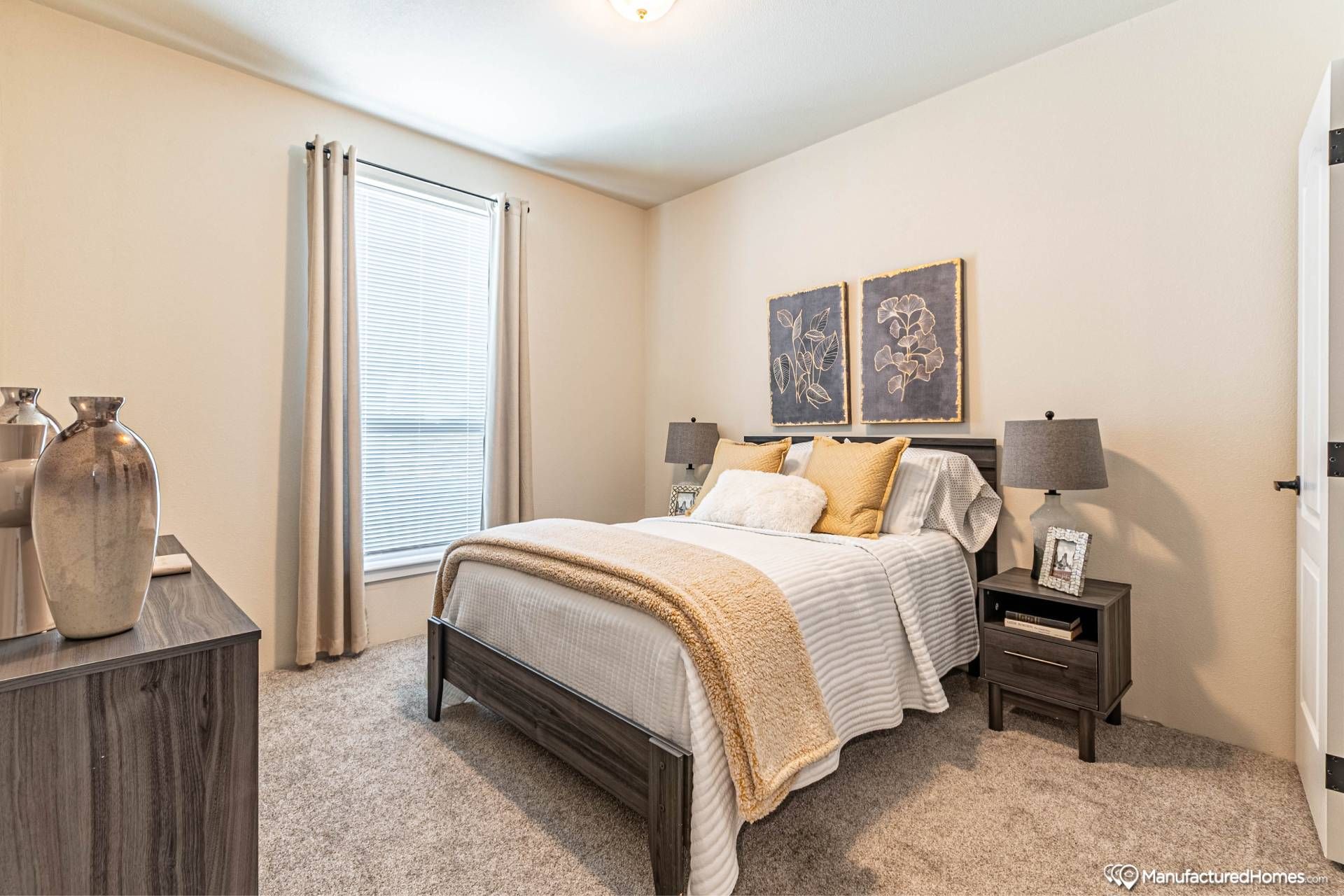 A cozy bedroom features a neatly made bed with white and beige linens, two bedside tables with lamps, and floral artwork on the wall. Soft light enters through a curtained window, creating a warm and inviting atmosphere.