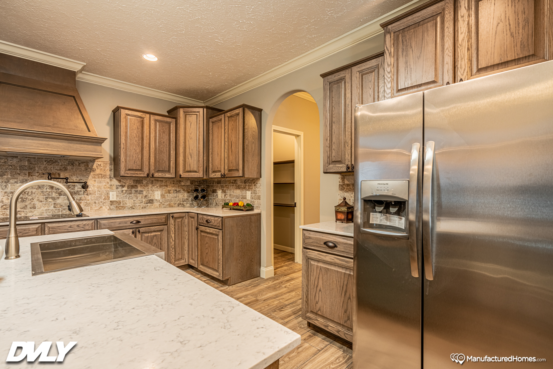 Warm, inviting kitchen with wooden cabinets, stainless steel fridge, marbled counter, and an arched doorway. Bright, clean, and modern setting.