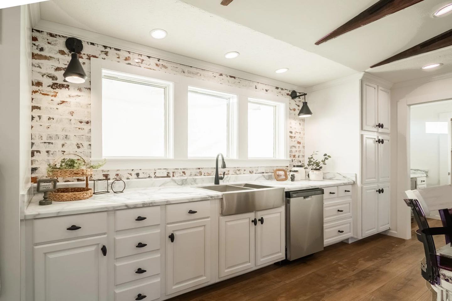 A bright kitchen with white cabinets, marble countertops, and a farmhouse sink. Exposed brick backsplash and wood floor create a rustic yet modern feel.