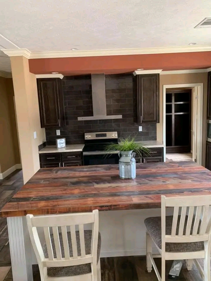 A modern kitchen with dark wooden cabinets and a black tile backsplash. A rustic wooden island features two chairs and a plant in a metal vase.