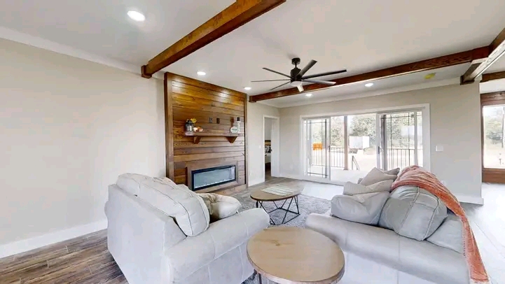 Spacious living room with beige sofas, wooden accent wall, modern fireplace, and large windows. Exposed beams and ceiling fan enhance cozy ambiance.