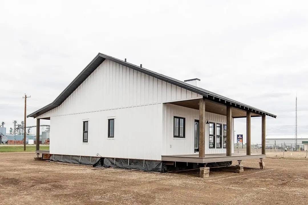 Single-story white house with a slanted roof and wooden porch, set on a dirt lot. The sky is overcast, creating a calm and neutral atmosphere.