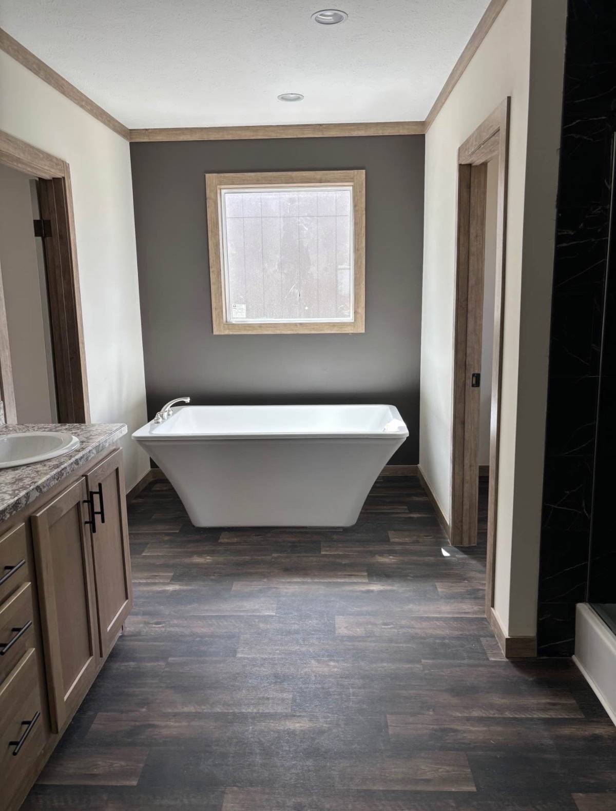 Modern bathroom with a free-standing white tub against a gray wall, framed by natural wood trim. Brown wooden floor and marble countertop create warmth.