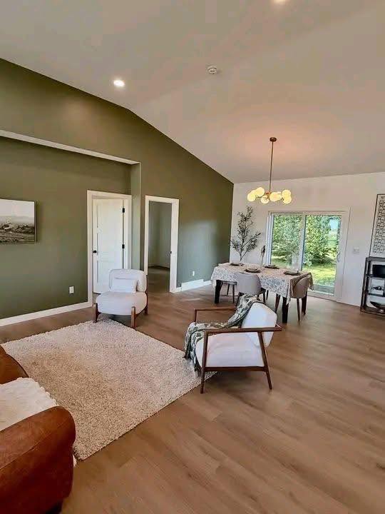 Spacious living room with green accent wall and vaulted ceiling. Cozy seating area on light wood floor, dining table with modern chandelier, large window showing greenery.