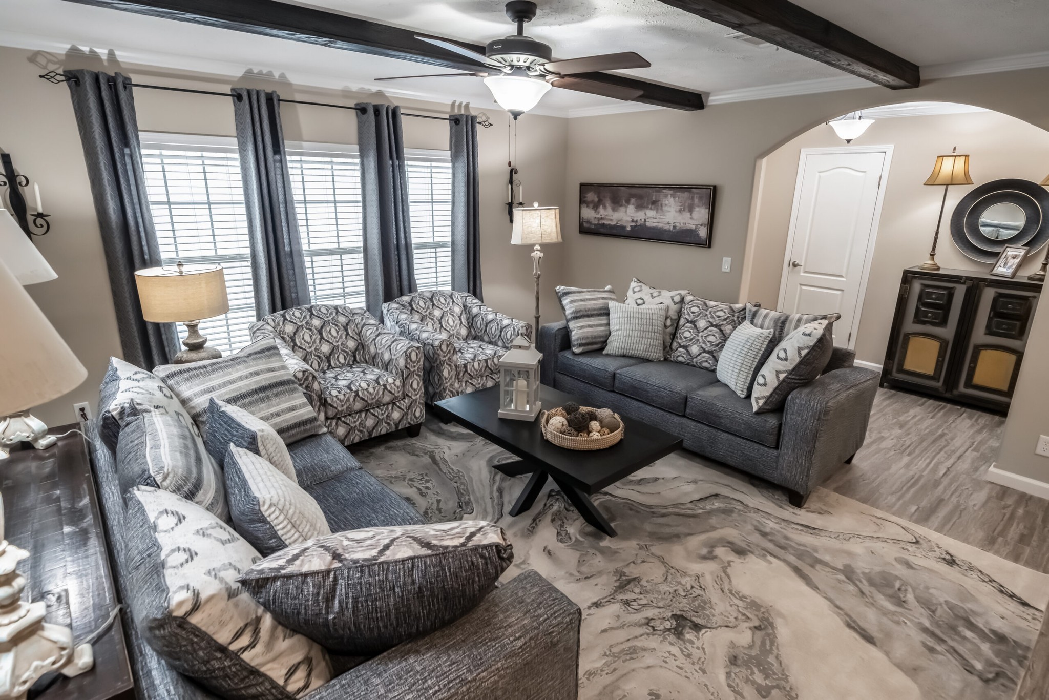 Stylish living room with gray sofas, patterned armchairs, and a modern coffee table. A ceiling fan, soft lighting, and a chic rug complete the cozy atmosphere.